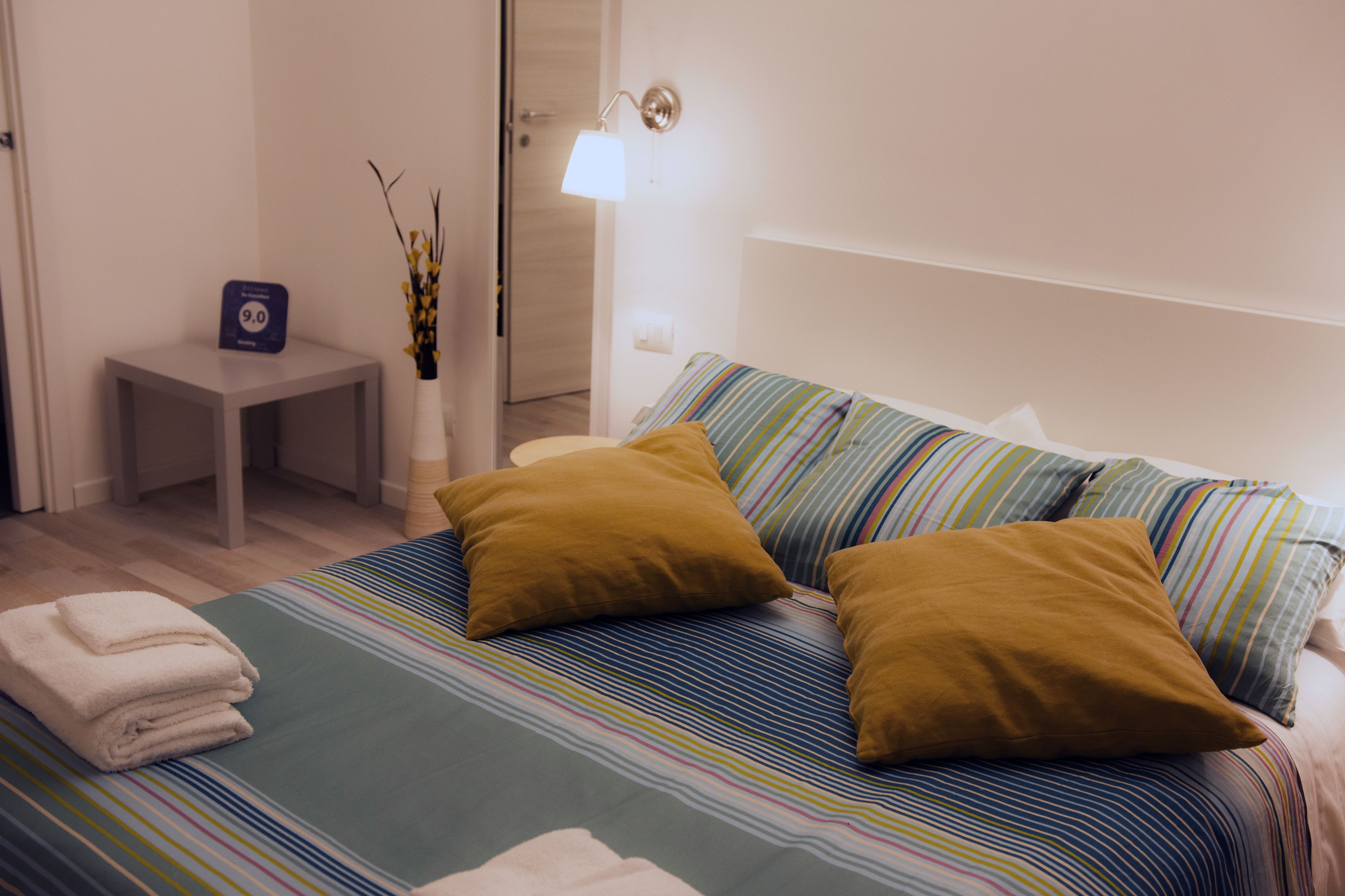 A neatly made bed with striped bedding and two pillows, a small bedside table with a lamp, and a vase with branches.