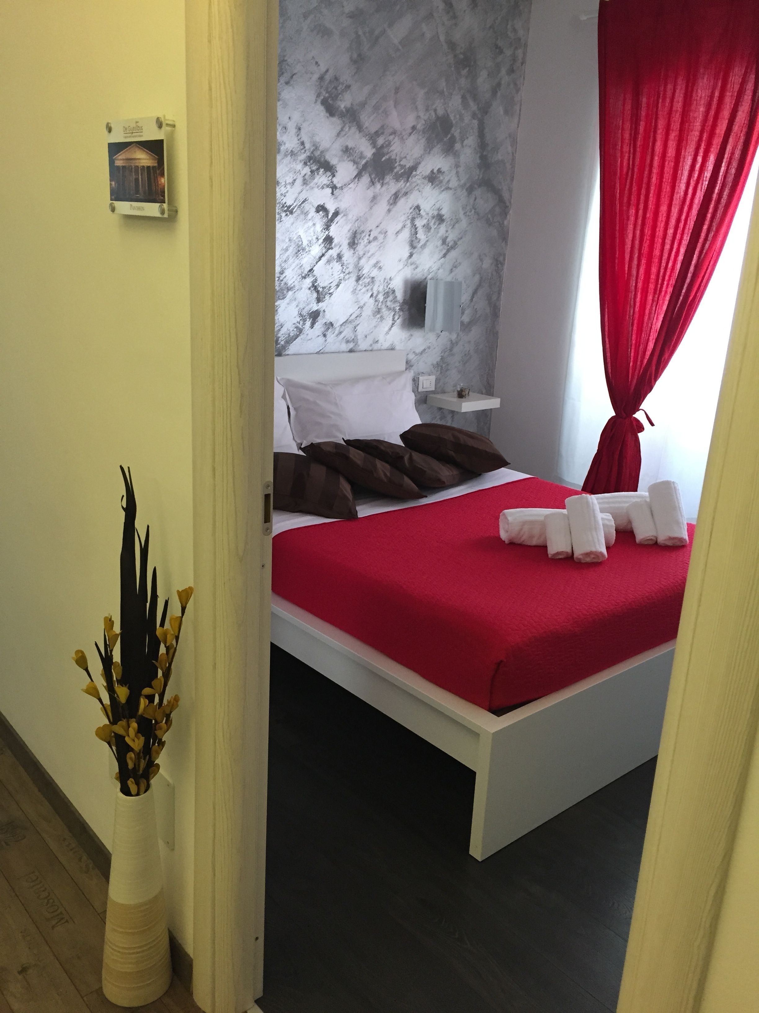 A bedroom with a bed, pillows, a nightstand, a vase with flowers, and a red curtain.