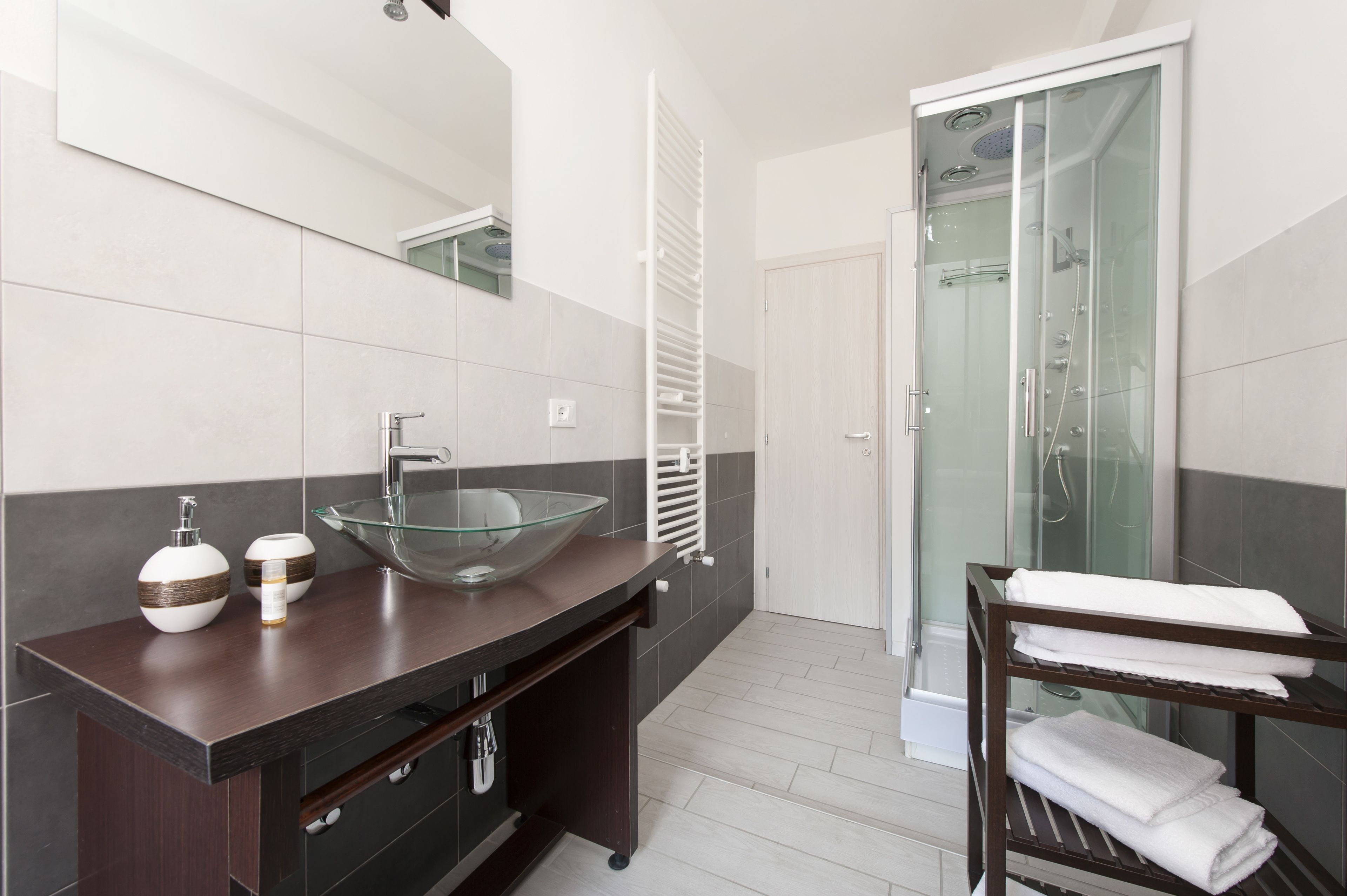 A modern bathroom with a glass bowl sink, dark wooden vanity, white tiles, a glass shower enclosure, and a towel rack.