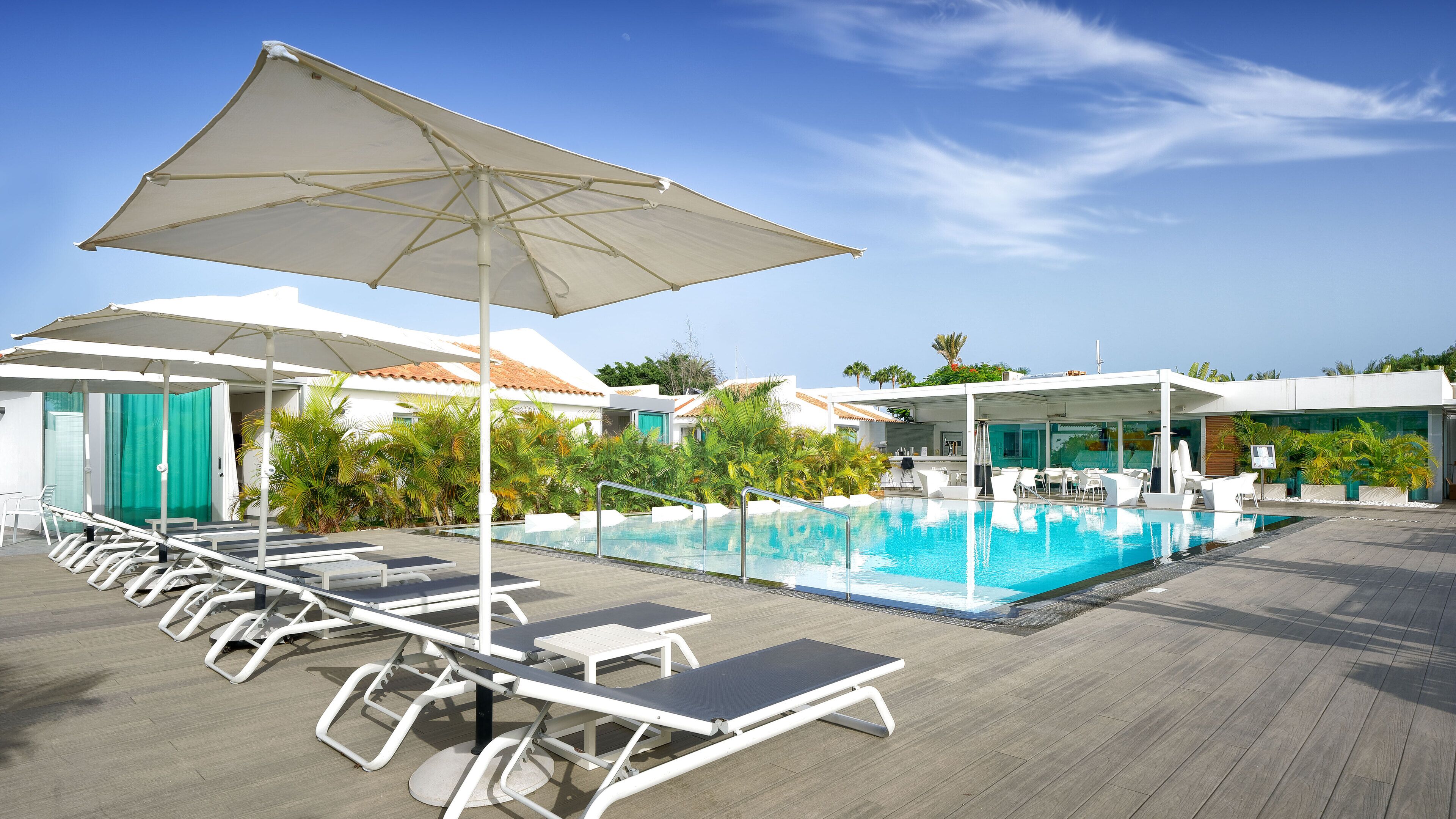 Un área junto a la piscina con sillones de descanso y sombrillas, un cielo azul despejado y una piscina.