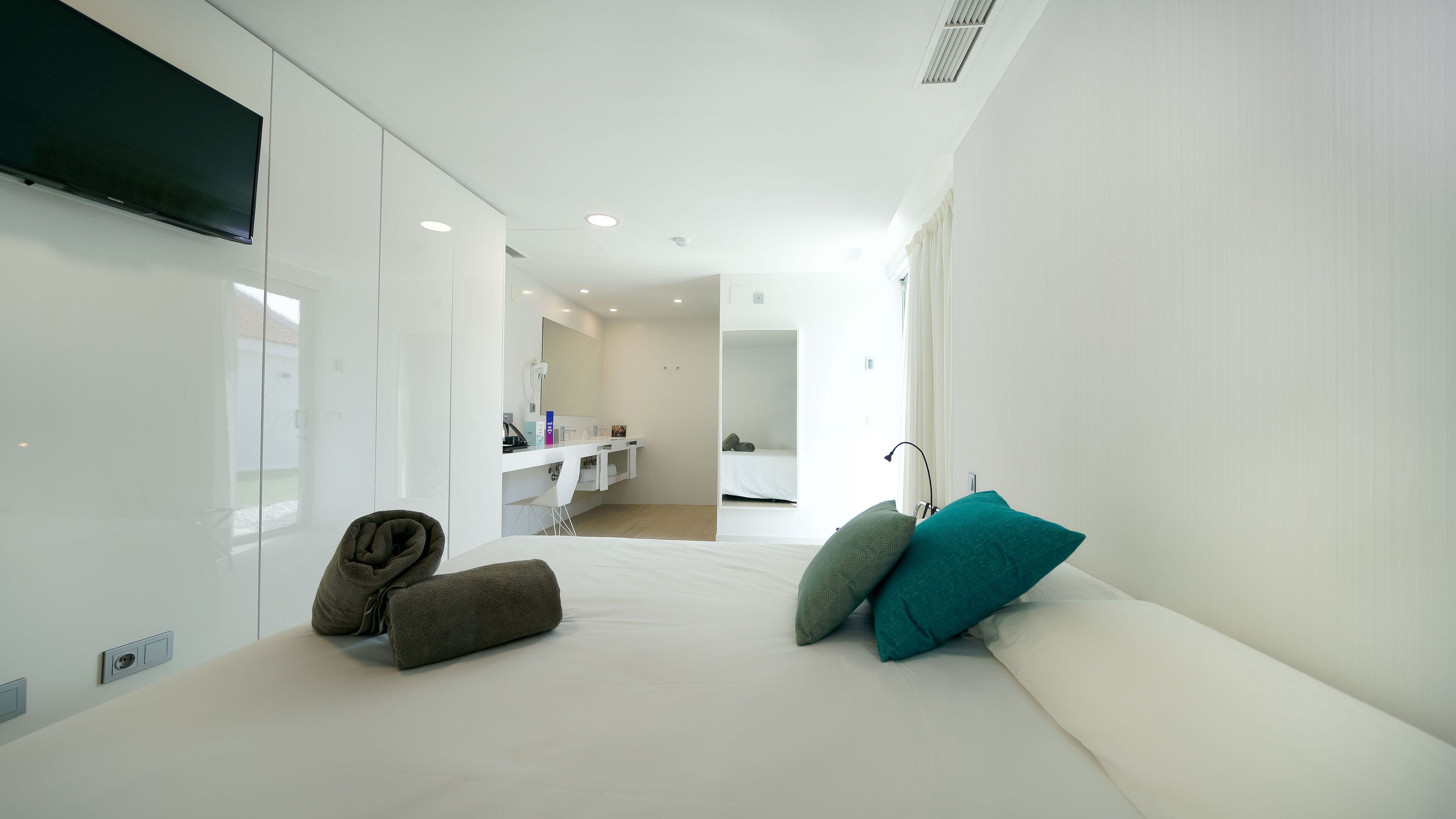 Een moderne hotelkamer met een groot bed, een aan de muur bevestigde televisie, een badkamer met spiegel en een klein zithoekje met kussens.