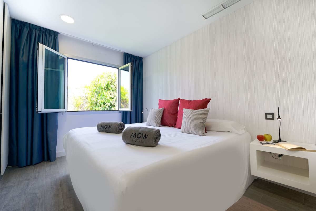 Ein modernes Hotelzimmer mit einem großen Bett, einem Nachttisch mit Lampe und Blick auf Grünflächen durch das Fenster.