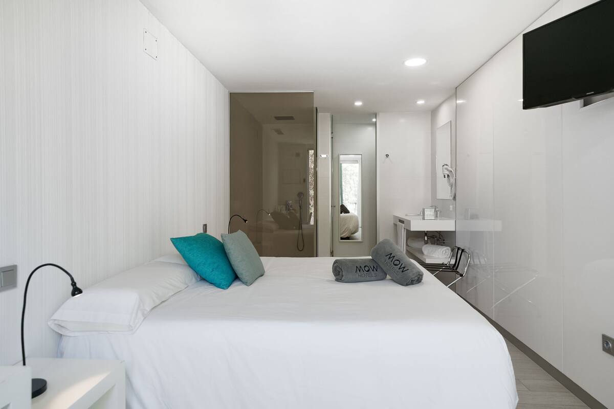 Ein modernes Hotelzimmer mit einem großen Bett, einem an der Wand montierten Fernseher, einem Badezimmer mit Spiegel und Waschbecken sowie einer Duschkabine.