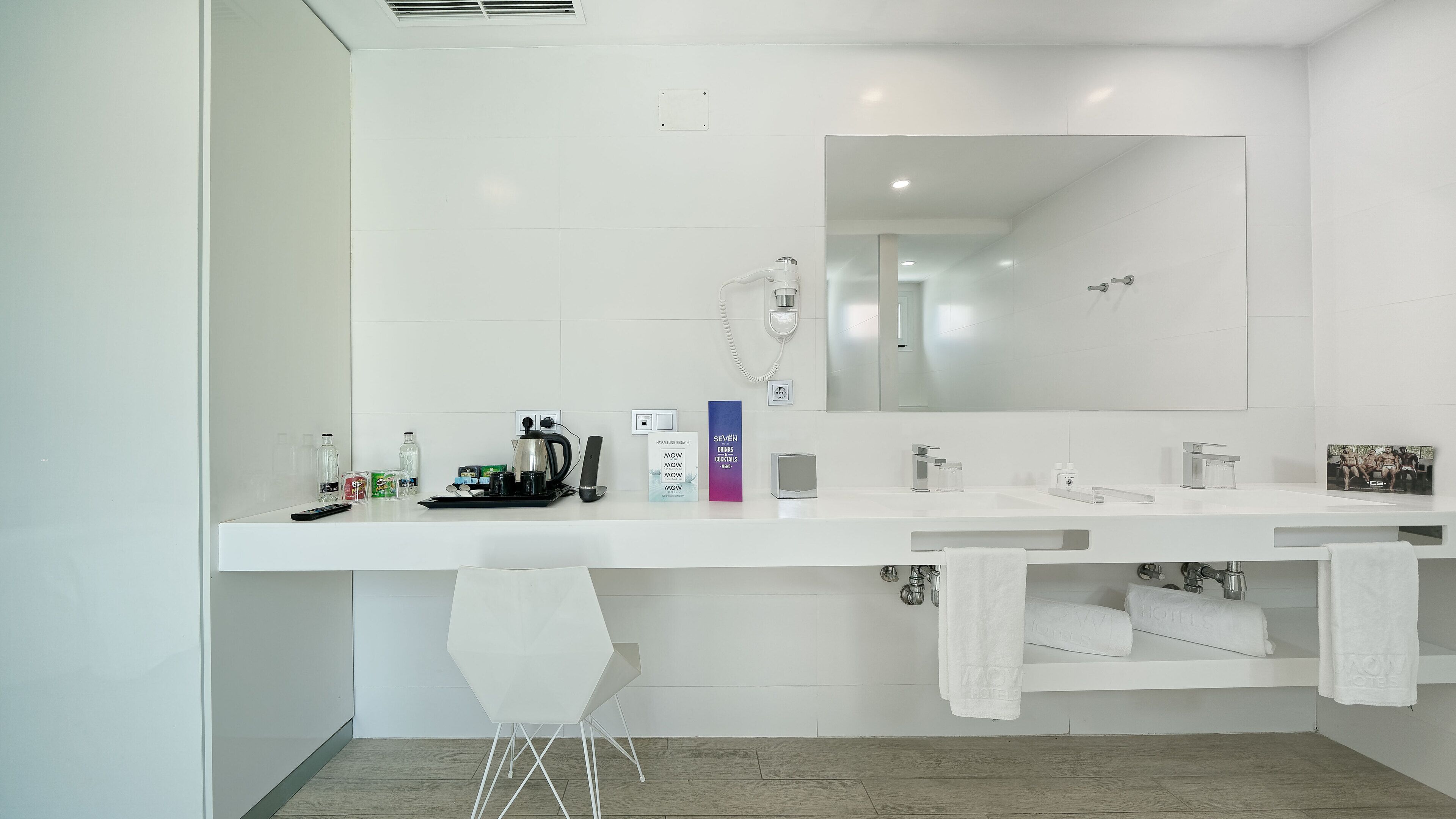 Un baño moderno con dos lavamanos, un espejo grande y una ducha. Hay artículos de aseo en el mostrador y un perchero con toallas.