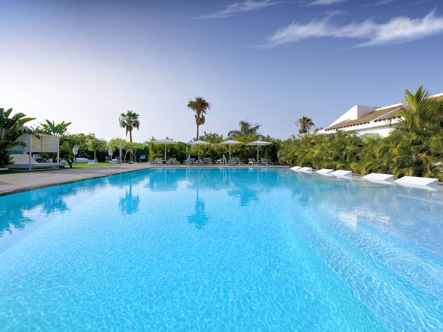 Una piscina azul cristalina con sillas de descanso y sombrillas, rodeada de palmeras y arquitectura moderna.