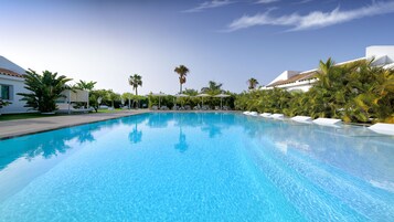 Una piscina azul cristalina con sillas de descanso y sombrillas, rodeada de palmeras y arquitectura moderna.