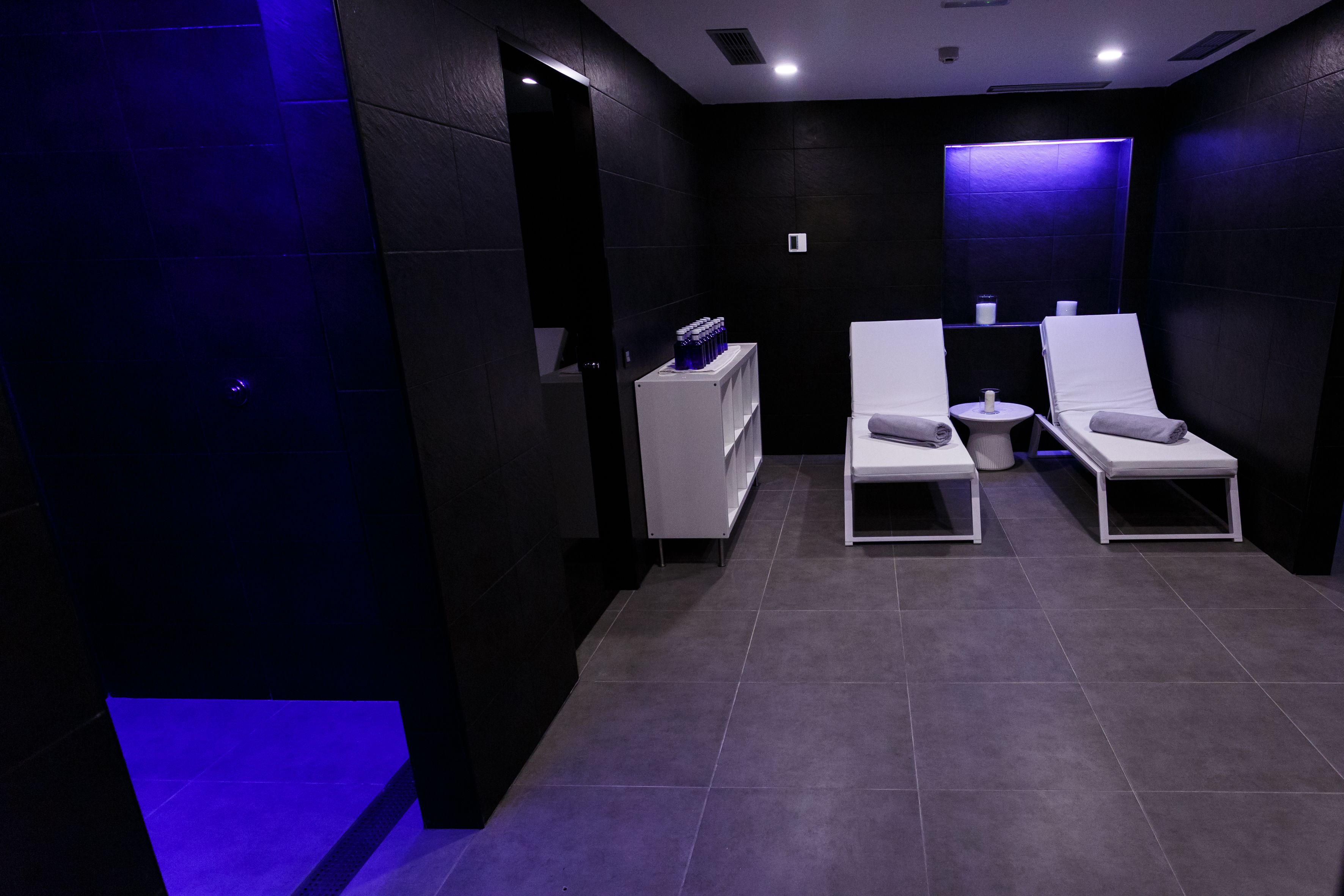 Une salle de spa moderne équipée de deux fauteuils inclinables, d’une petite table blanche avec une serviette et d’une étagère contenant des bouteilles.