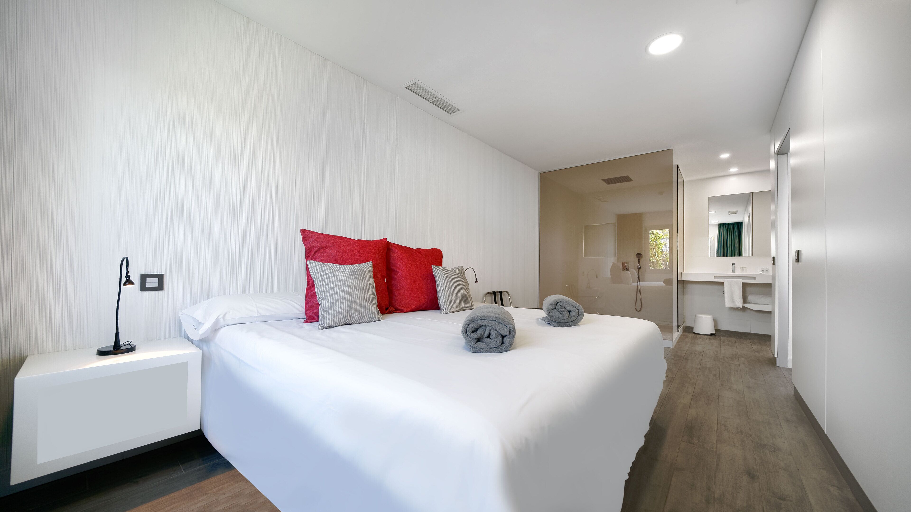 A modern hotel room with a neatly made bed, a bedside table with a lamp, and a bathroom visible through an open door.