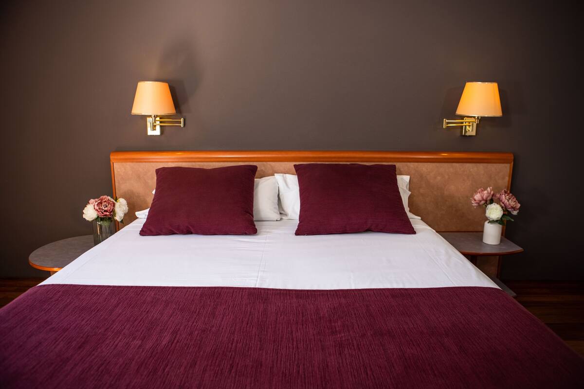 Cama bem arrumada com cabeceira de madeira, dois travesseiros e um cobertor vermelho. Há duas mesas de cabeceira com luminárias e vasos de flores.