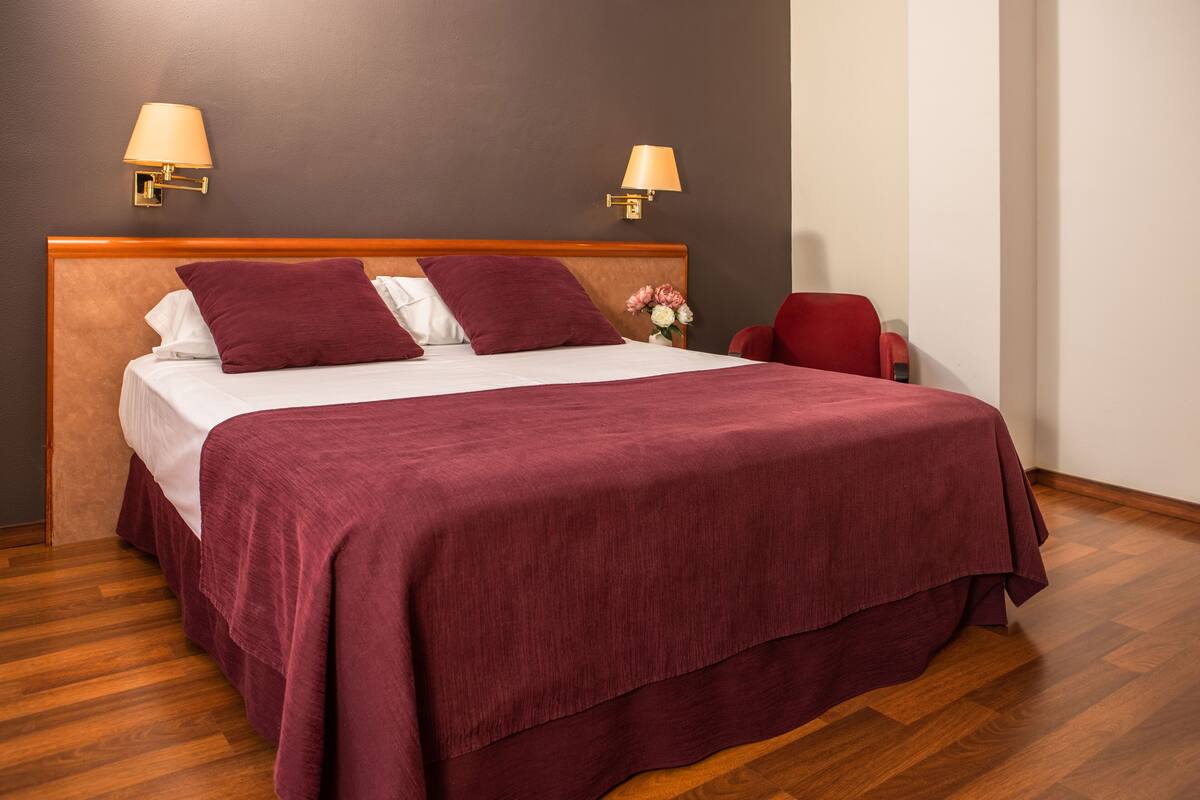 Uma cama bem arrumada com colcha e travesseiros vermelhos, duas luminárias fixadas na parede e uma cabeceira de madeira.