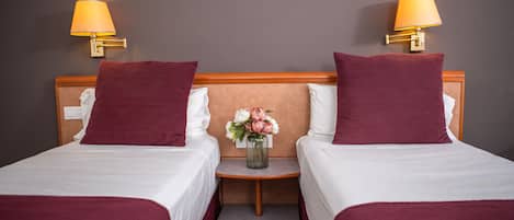 A hotel room with two single beds, each with a red blanket, white pillows, and a small nightstand with a vase of flowers.