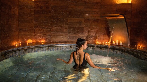 A woman in a black swimsuit in a stone-walled indoor pool with a waterfall feature and candles. - RVHotels Spa Vila de Caldes (Caldes de Montbui)