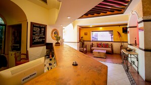 A hotel lobby with a reception desk, a seating area with a sofa and chairs, a wall clock, and a menu board listing room rates.
