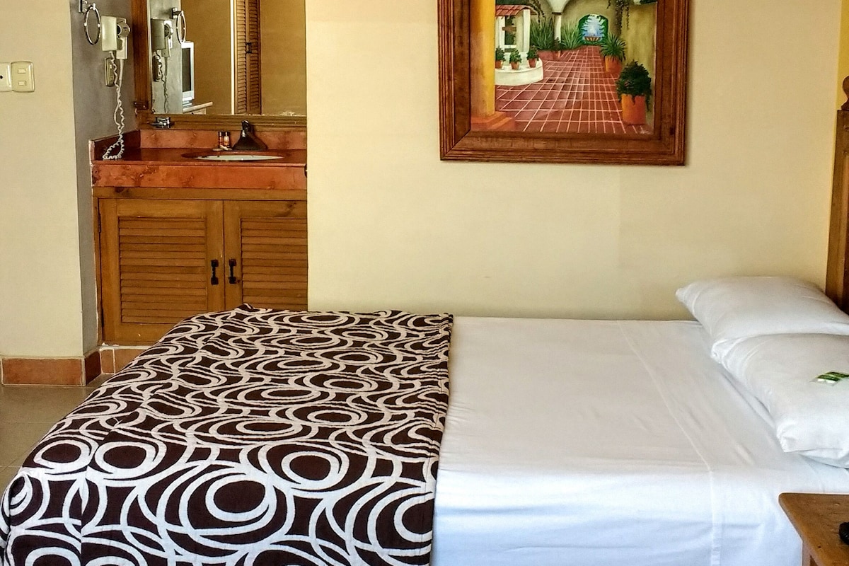 A hotel room with a bed, a large mirror, a painting of a landscape, and a wooden vanity with a sink.