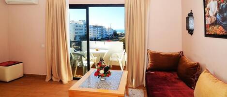 A living room with a sofa, a coffee table with a vase of flowers, a balcony with a view of buildings, and a painting on the wall.