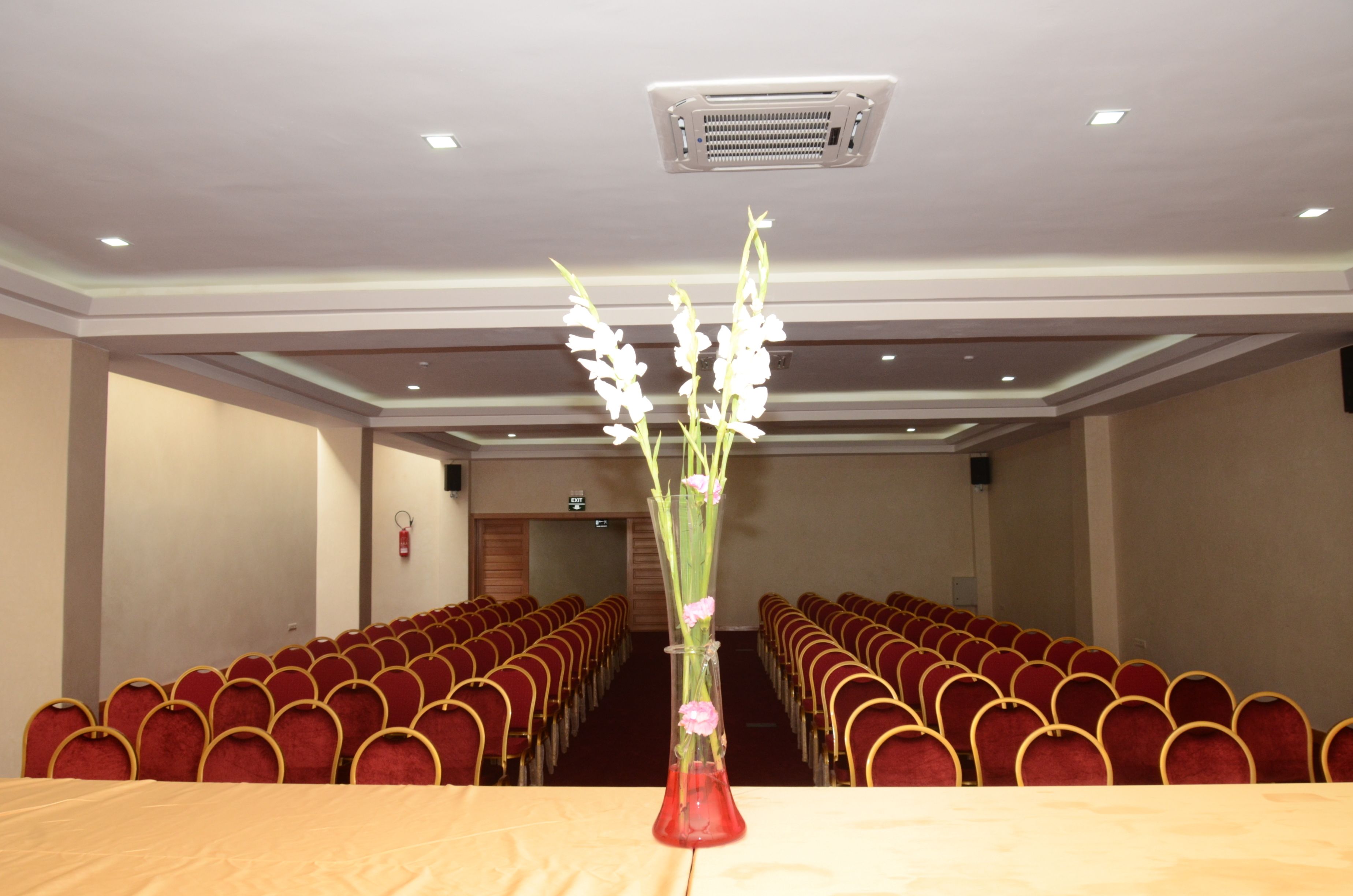 Ein Konferenzraum mit Reihen roter Stühle, einem langen Tisch und einer Blumenvase.