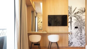 A room with a wooden wall, a mounted TV, two white chairs, a small table, and a decorative wall with palm tree and flower patterns.
