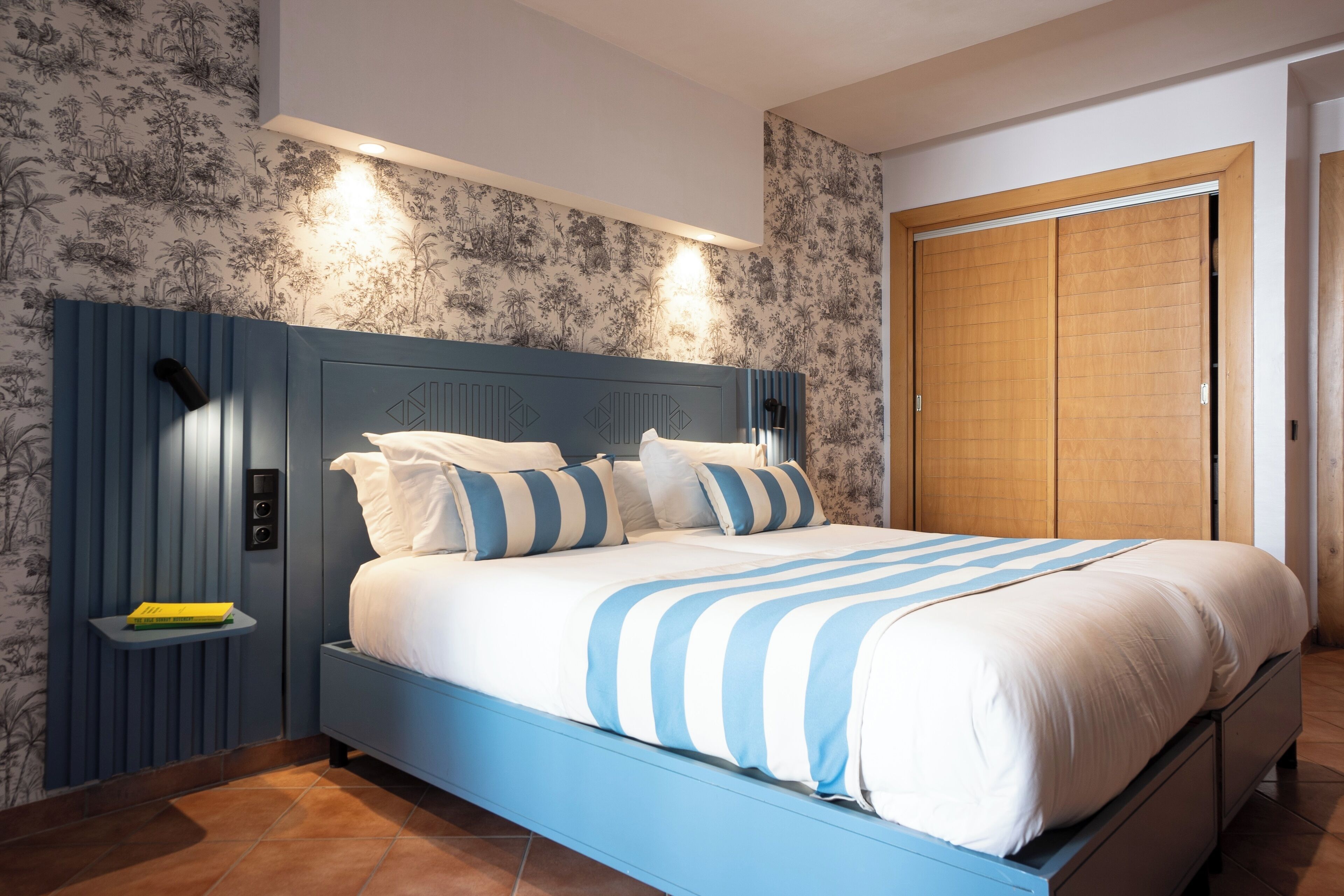 Un dormitorio con cabecera azul, cama con ropa de cama a rayas blancas y azules, mesita de noche con un libro y un armario de madera.