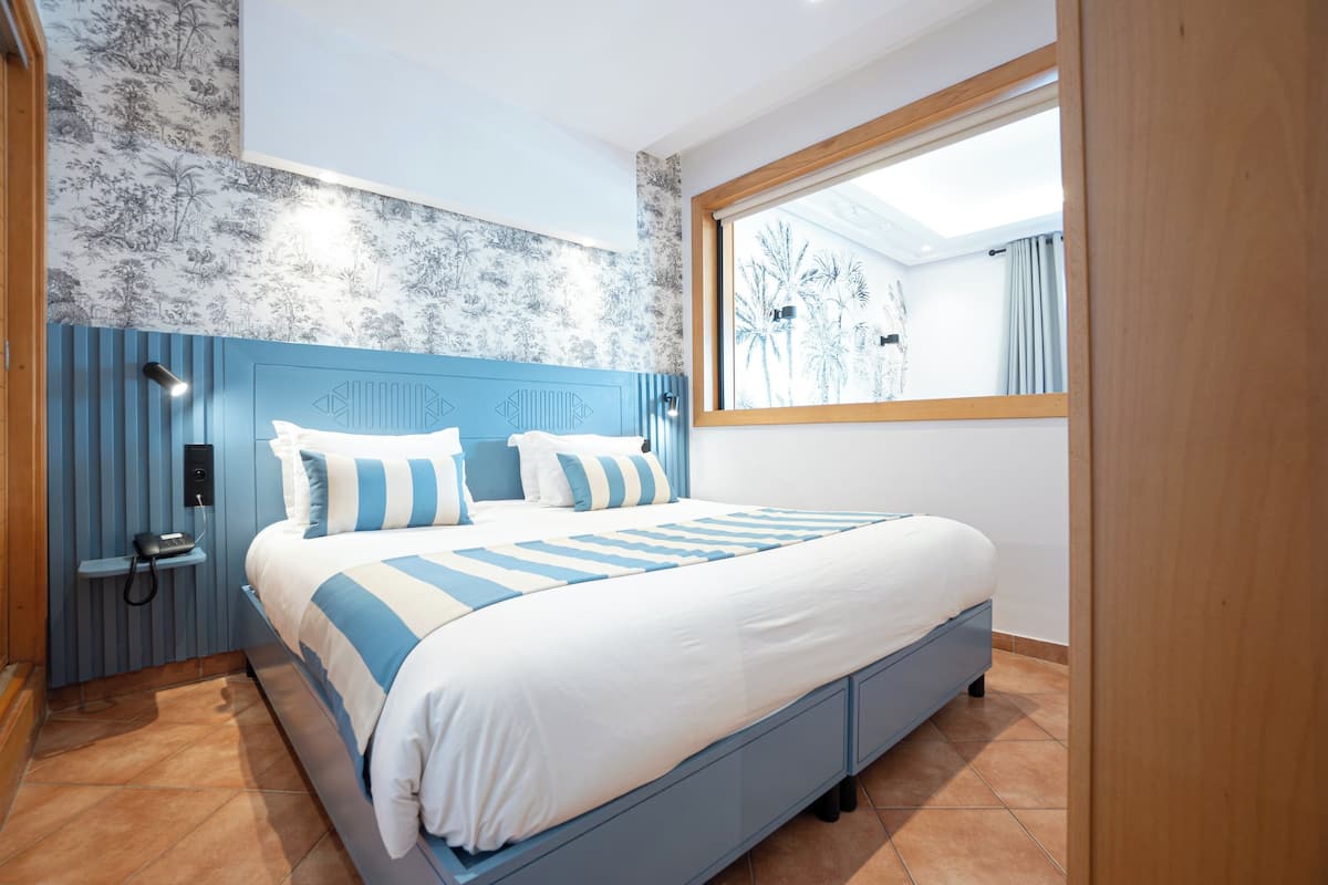 A bedroom with a bed, a nightstand, a telephone, and a window with a view of palm trees.