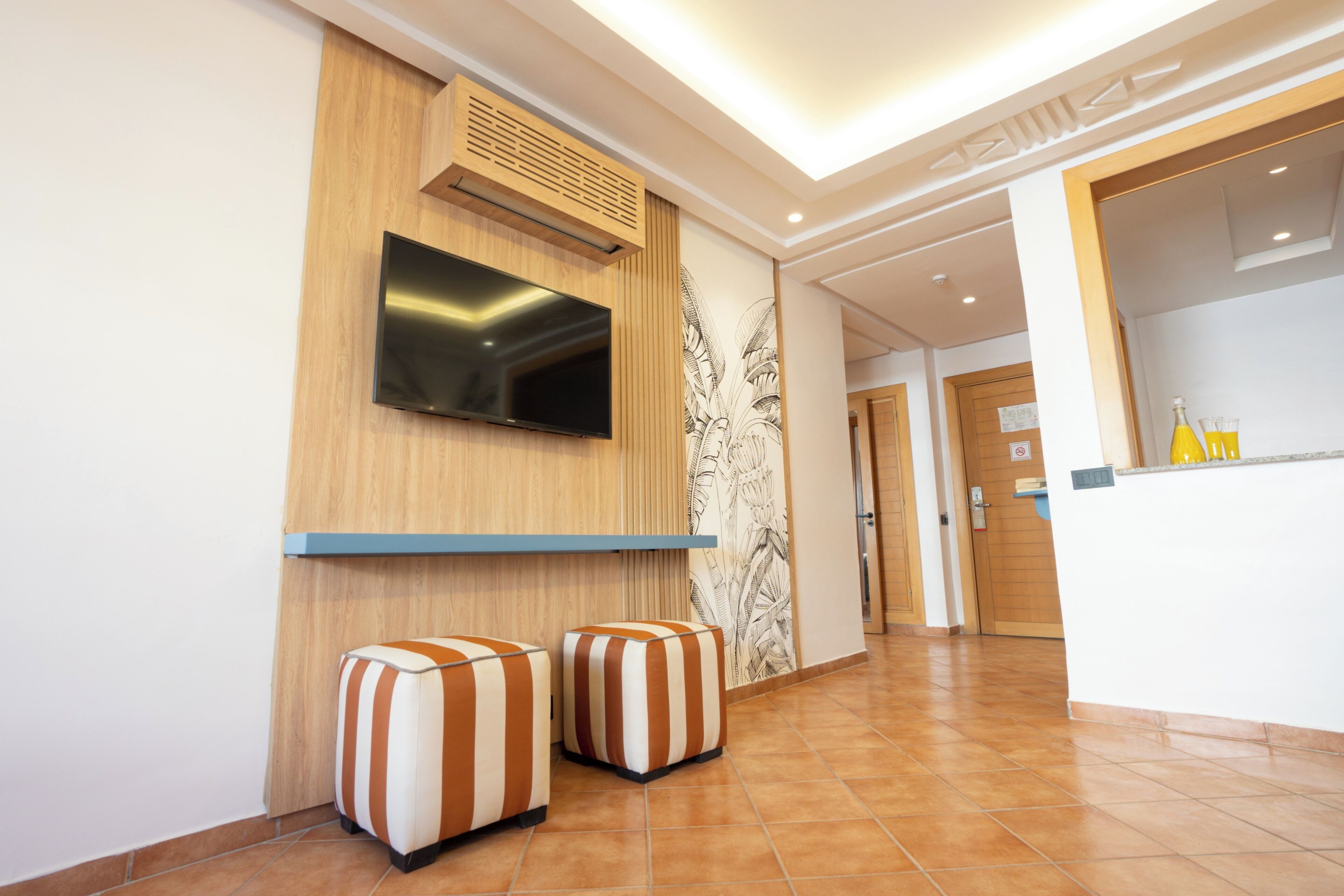 Un moderno vestíbulo de hotel con una televisión de pantalla plana montada en una pared de madera, dos otomanos a rayas y un mostrador de recepción.