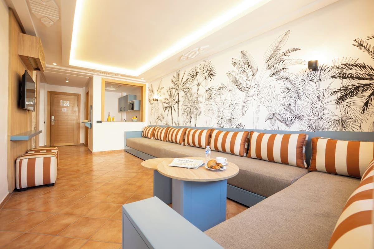A modern hotel lobby with a sofa, striped cushions, a round table with a menu, and a wall with a tropical leaf pattern.