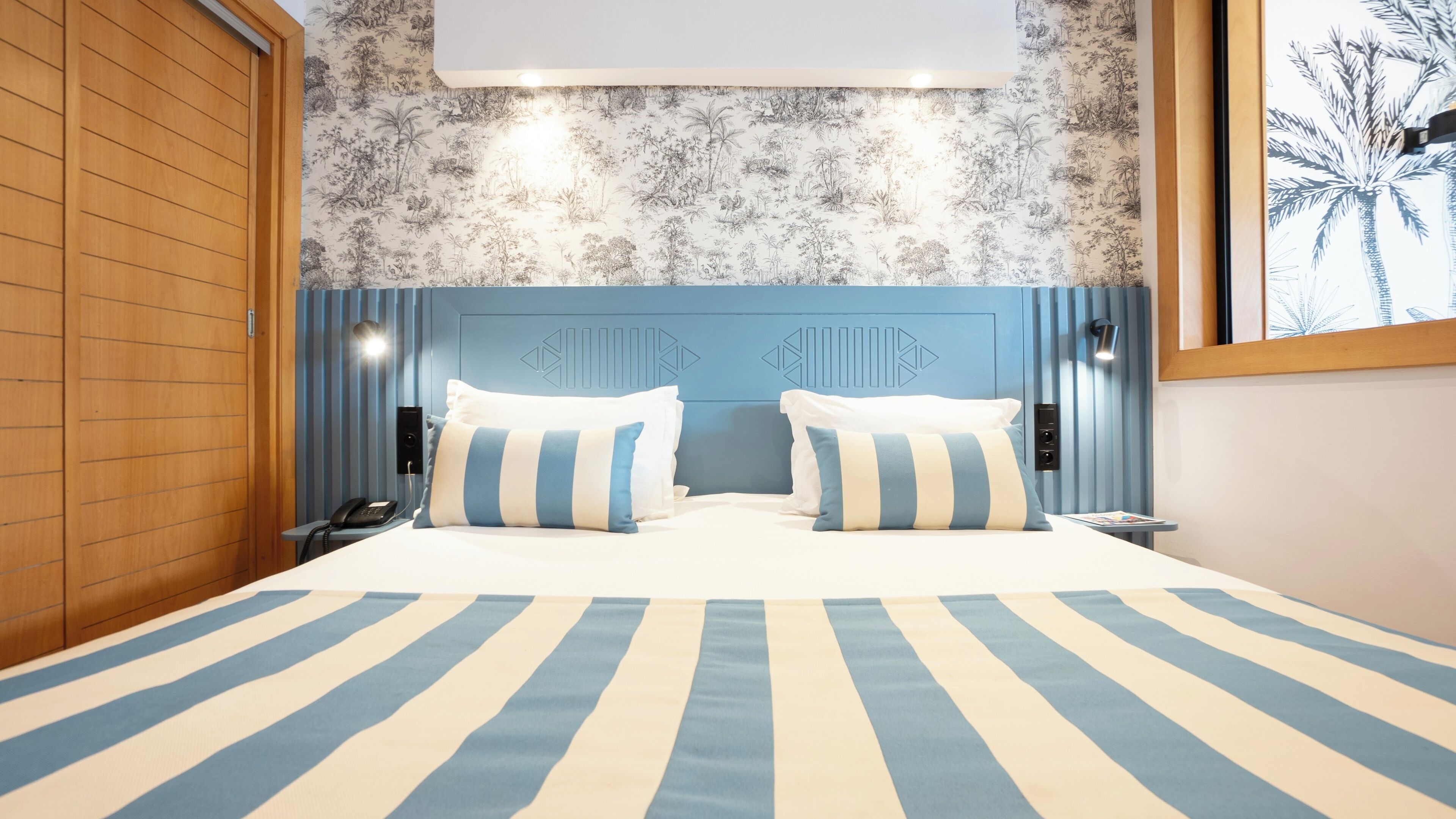 A hotel room with a large bed, striped bedding, a wooden wardrobe, a wall with a tropical mural, and a nightstand with a phone and lamp.