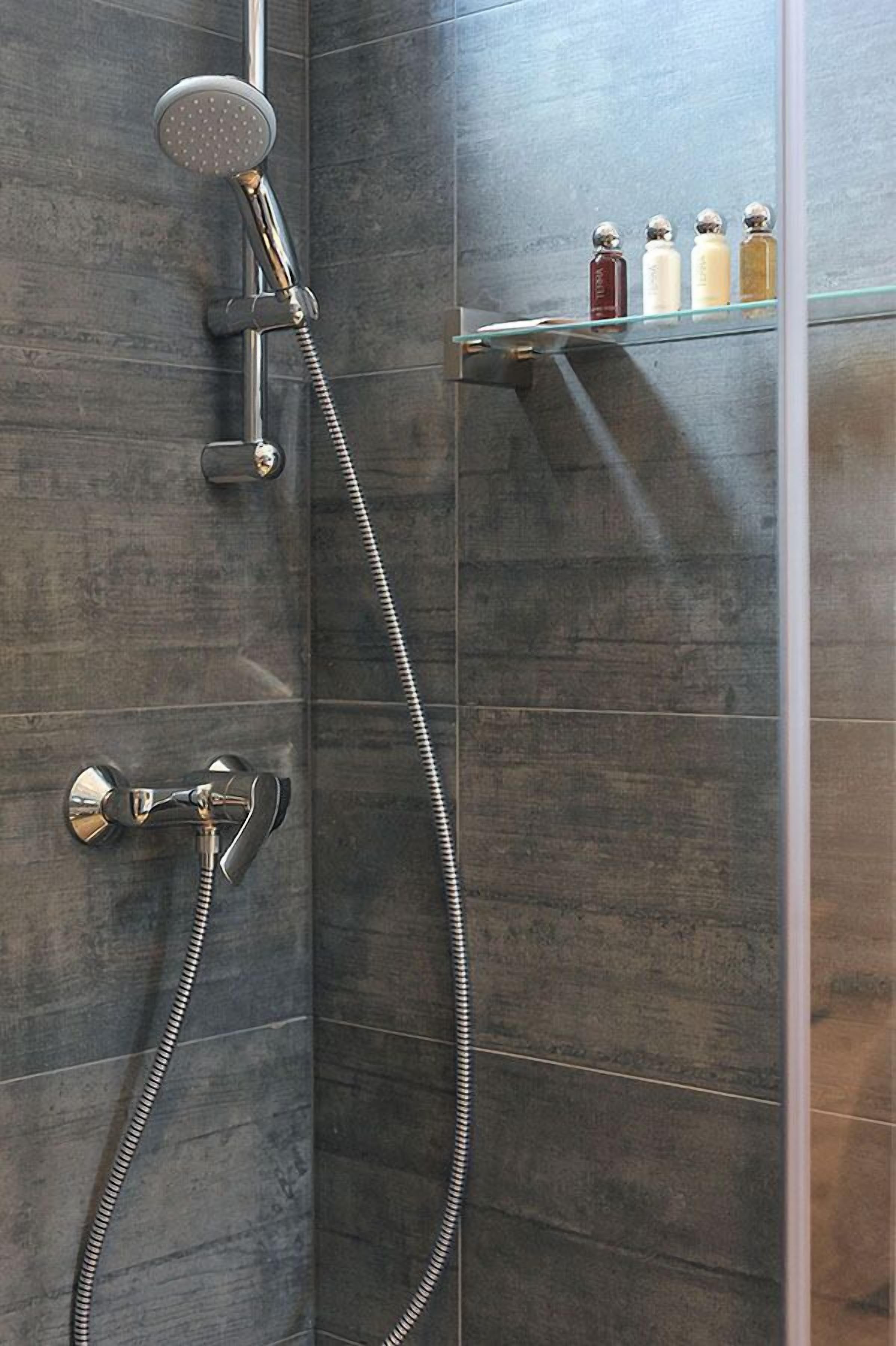 Een douche met een handdouche en flexibele slang, een kraan op de muur en een glazen plank met toiletartikelen.