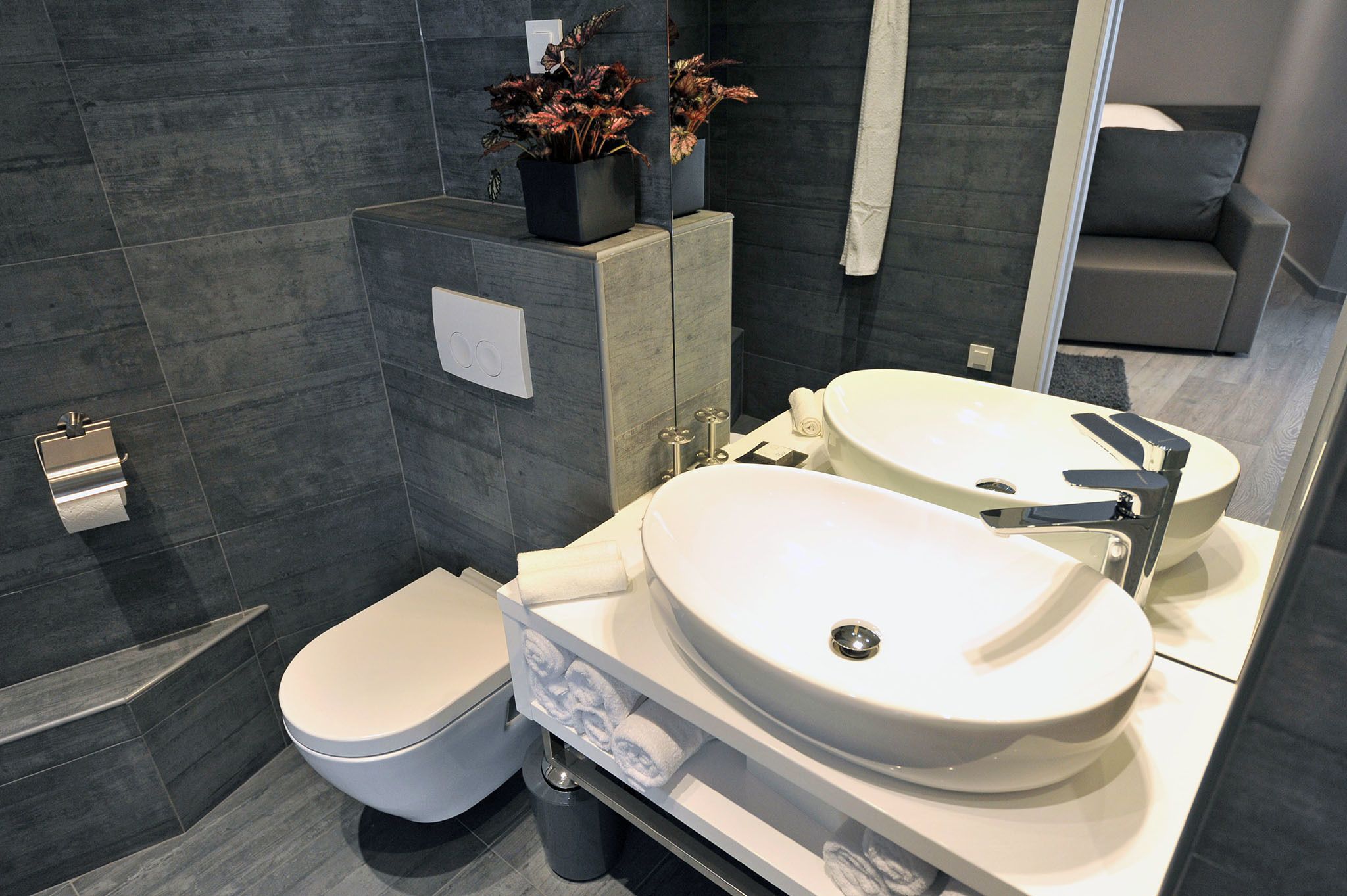 A modern bathroom with a toilet, two sinks, a mirror, and a potted plant.