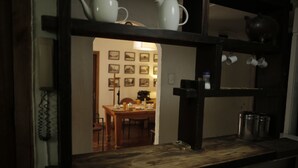 A kitchen with wooden shelves holding white mugs and a kettle. There is a dining area in the background with a table and chairs. - Hostal ByBSiglo (Santiago)