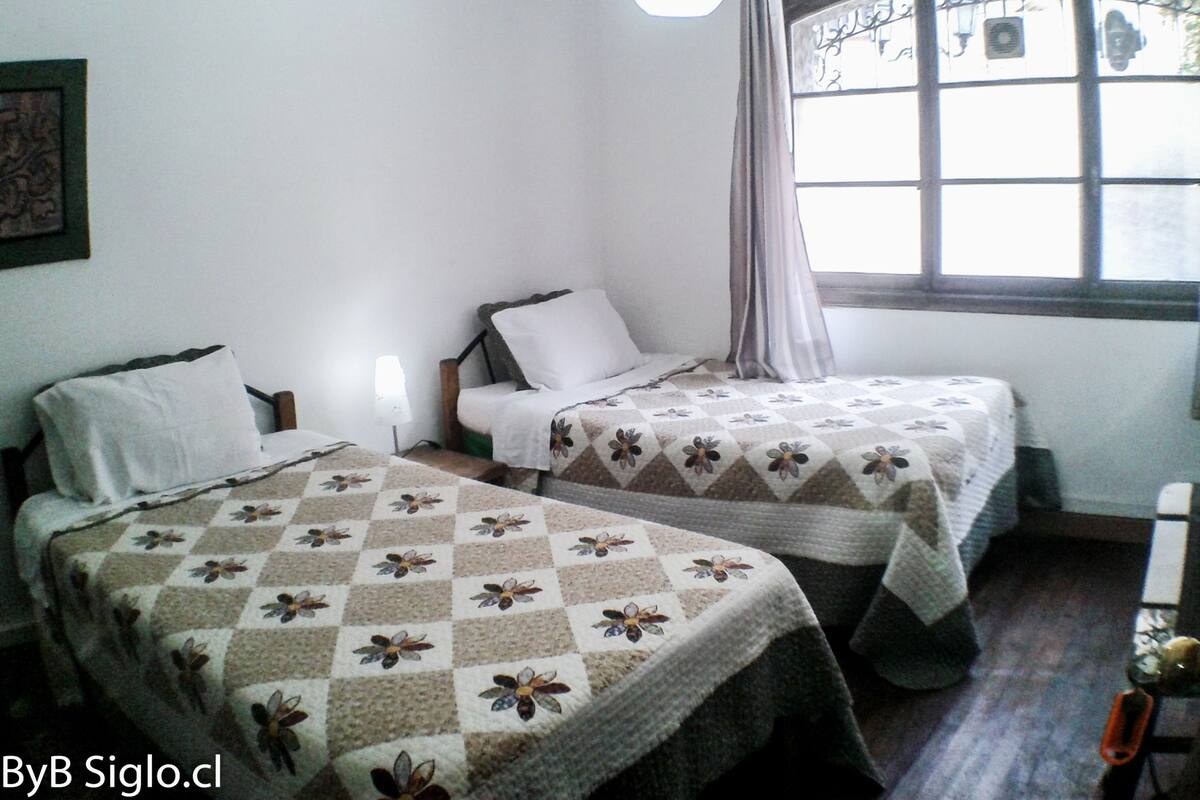A room with two beds, each with a floral-patterned quilt, a bedside table with a lamp, and a window with curtains.