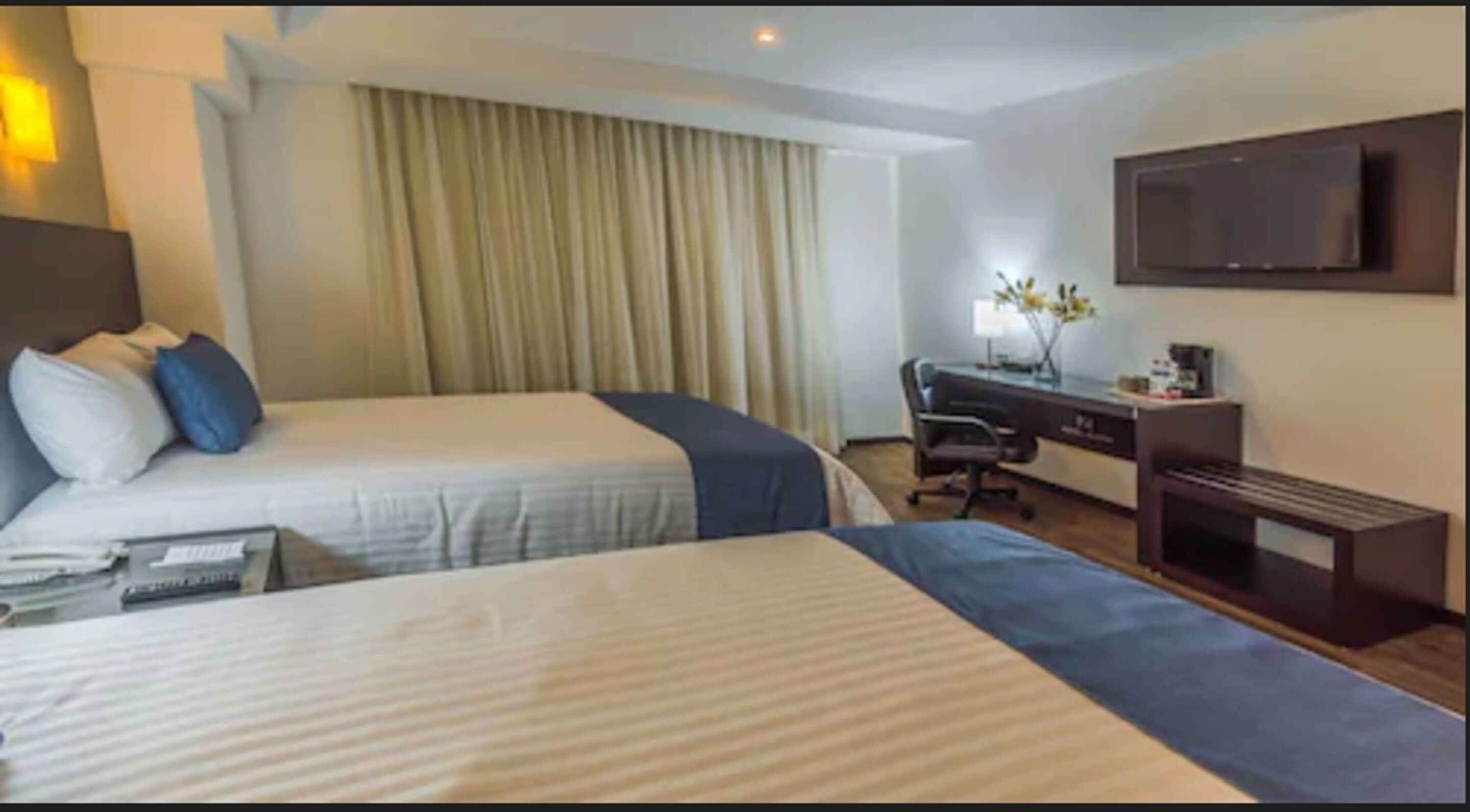 A hotel room with two beds, a desk, a chair, a TV, and a window with curtains.