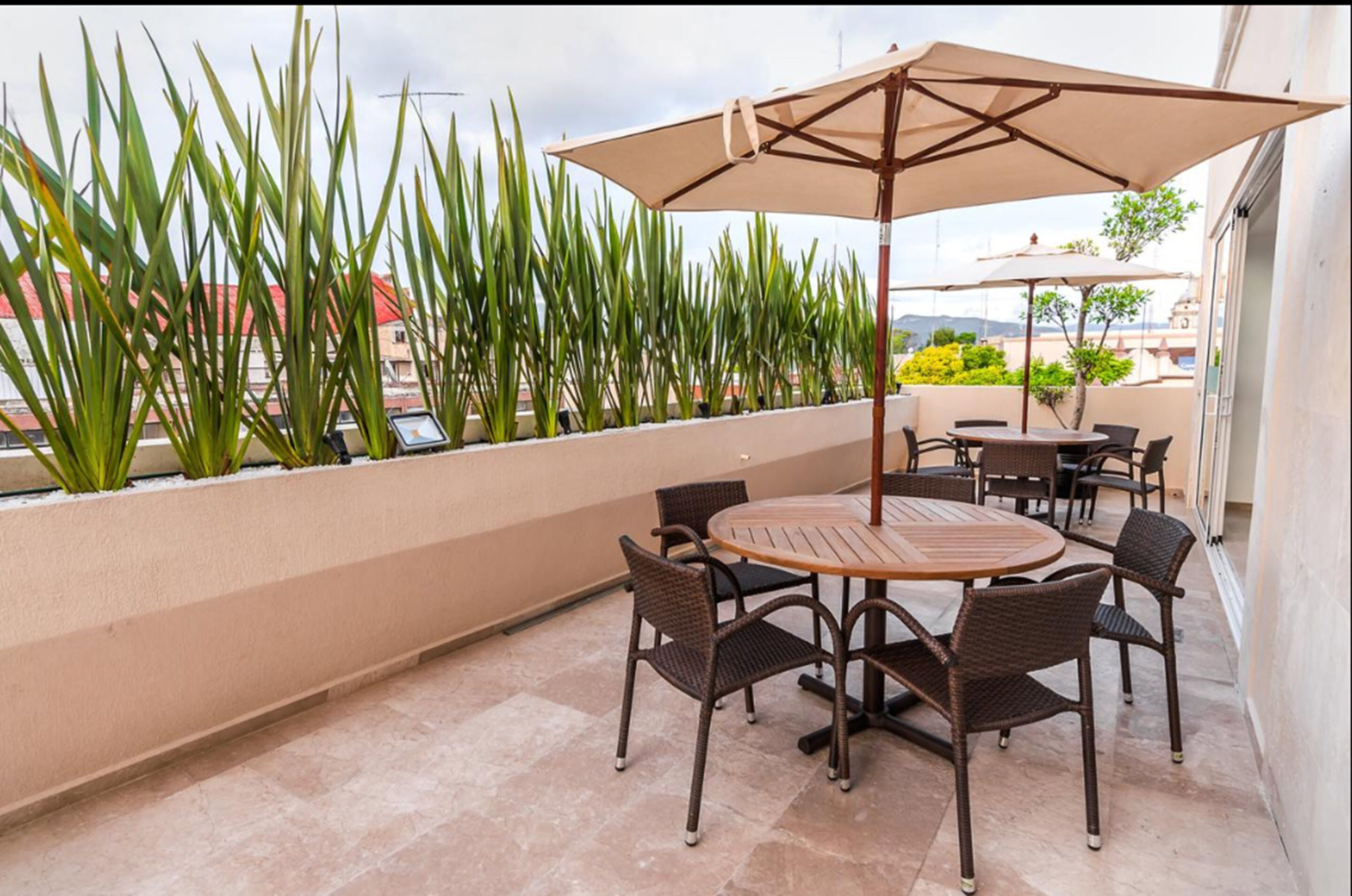 Un patio con una mesa redonda y sillas, rodeado de plantas altas y un paraguas grande.