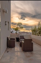 Une terrasse sur le toit, aménagée avec des meubles en osier et un arbre en pot, d’où l’on peut admirer le panorama urbain au coucher du soleil.