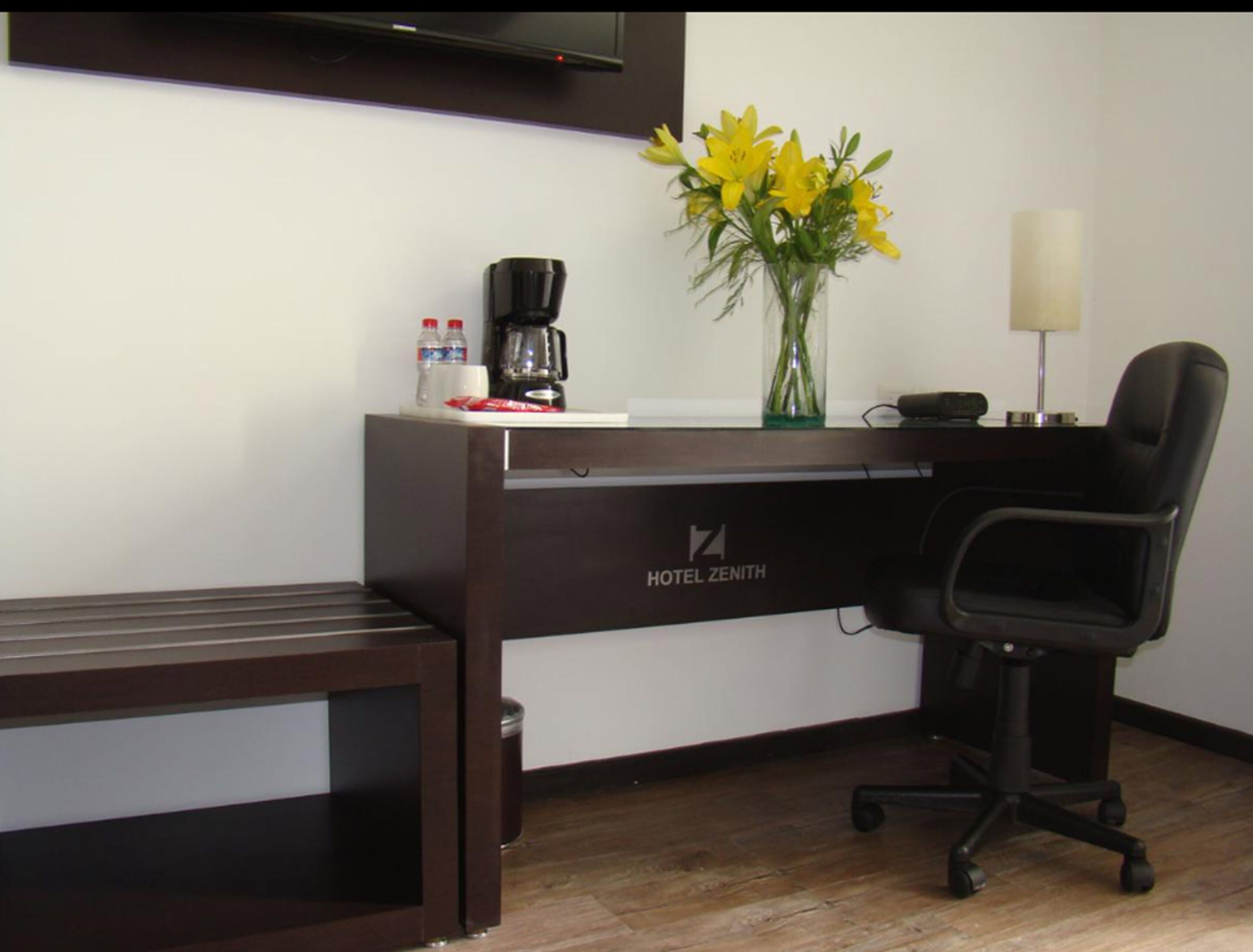 A hotel desk with a coffee maker, water bottles, a vase of yellow flowers, a lamp, and a black office chair.