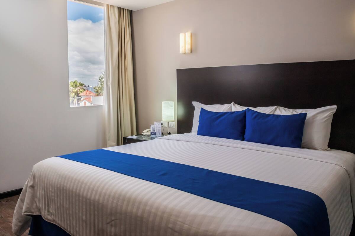 Una camera d'albergo con un letto grande, un comodino con telefono e lampada, una finestra con tende e vista sugli edifici esterni.