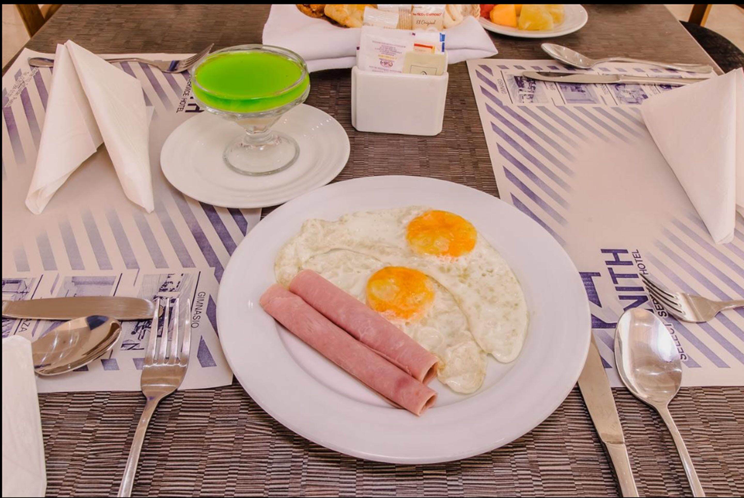 Un plato de desayuno con huevos, jamón y una bebida verde sobre una mesa con un camino de mesa a rayas.