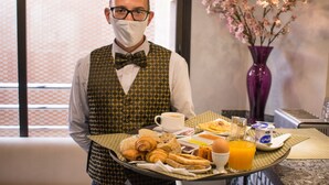 A breakfast tray with croissants, eggs, and orange juice, held by a person in a patterned vest and bow tie.