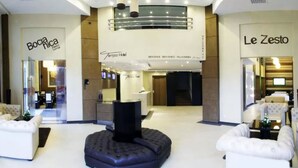 A modern hotel lobby with a reception desk, seating area, and signs for Boccia Rica restaurant and Le Zesto.