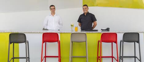 Two men standing behind a counter with a drink and a phone. There are four chairs in front of the counter.
