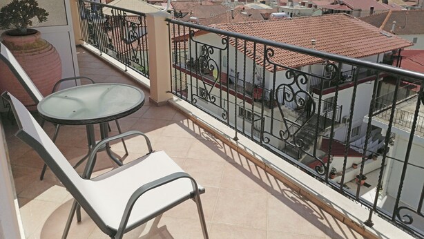 A balcony with a glass table and two chairs, overlooking a coastal town with terracotta rooftops. - Villa Maria (Parga)