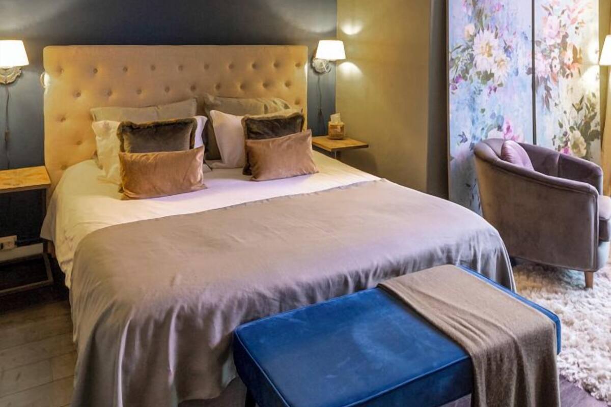 A bedroom with a large bed, two bedside tables, a chair, a bench, and a chandelier.