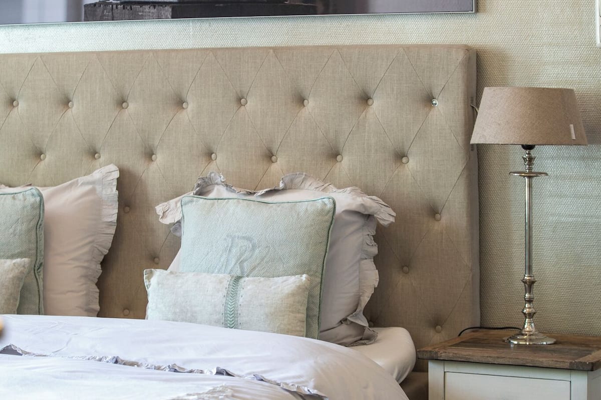 A bedroom with a tufted headboard, a bed with white linens, a bedside table with a lamp, and a wall-mounted artwork above the bed.