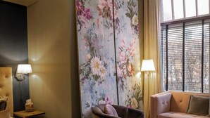 Habitación de hotel con cama, sofá, una silla, un cuadro grande con motivos florales en la pared y una ventana con persianas.