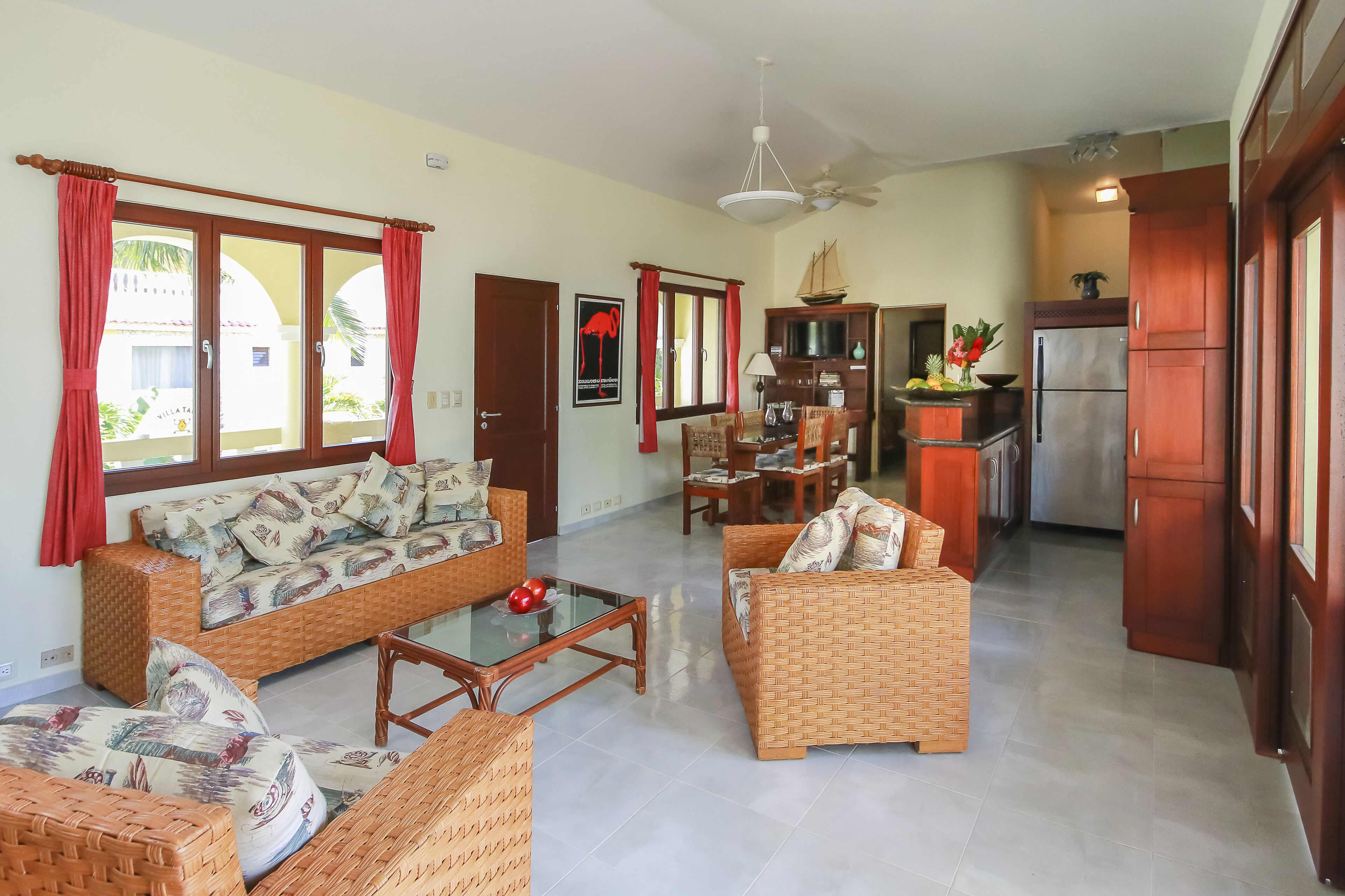 A spacious living area with wicker furniture, a glass coffee table, and a kitchen with wooden cabinets and a refrigerator.