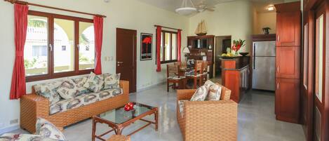 A spacious living area with wicker furniture, a glass coffee table, and a kitchen with wooden cabinets and a refrigerator.