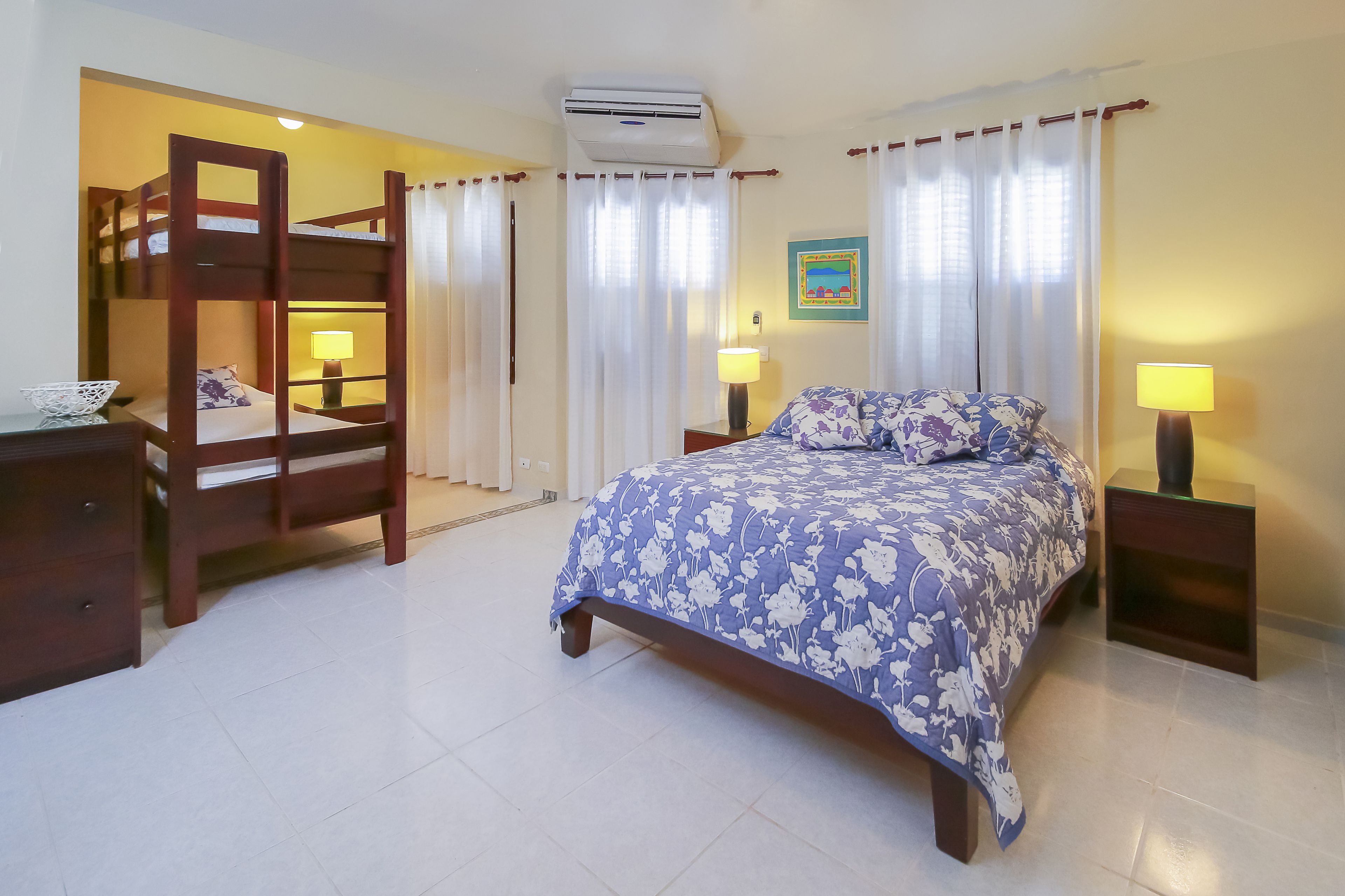 A bedroom with a bunk bed, a single bed with a floral bedspread, two bedside tables with lamps, a wall-mounted air conditioner, and a framed picture on the wall.