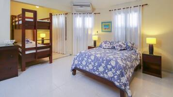 A bedroom with a bunk bed, a single bed with a floral bedspread, two bedside tables with lamps, a wall-mounted air conditioner, and a framed picture on the wall.