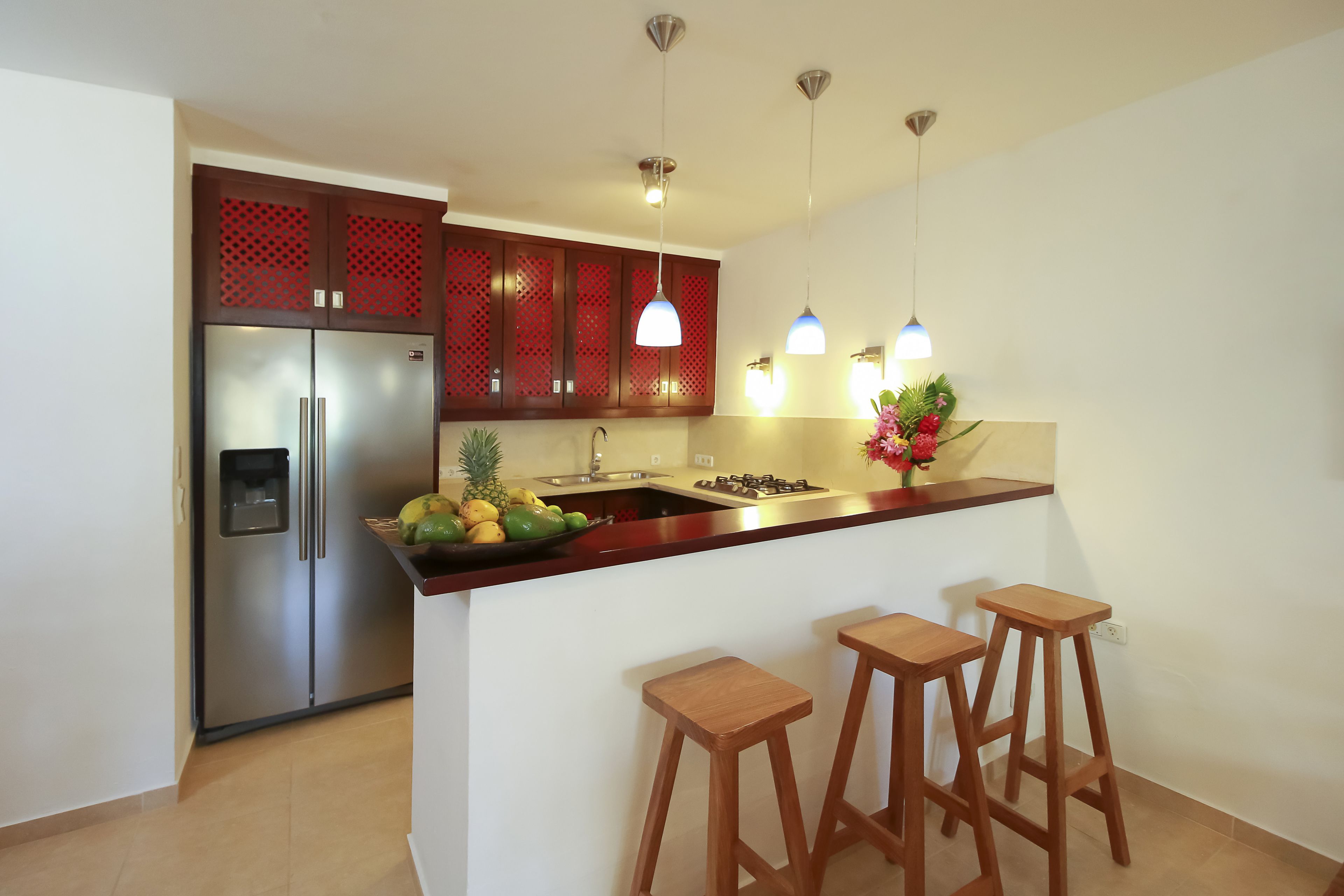 A modern kitchen with a stainless steel refrigerator, wooden bar stools, and pendant lighting.