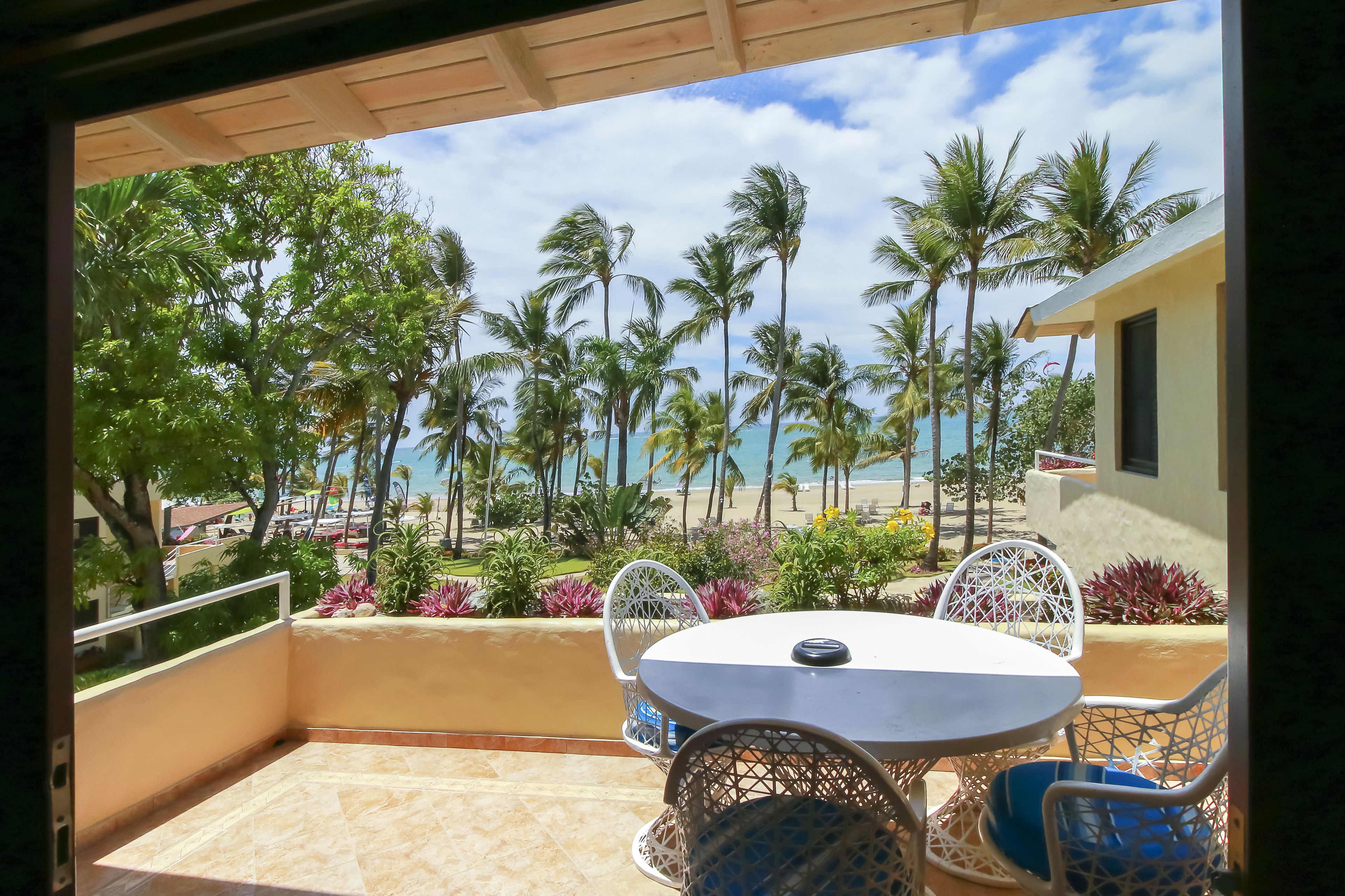 Een balkon met een tafel en stoelen, met uitzicht op een strand met palmbomen en een heldere lucht.