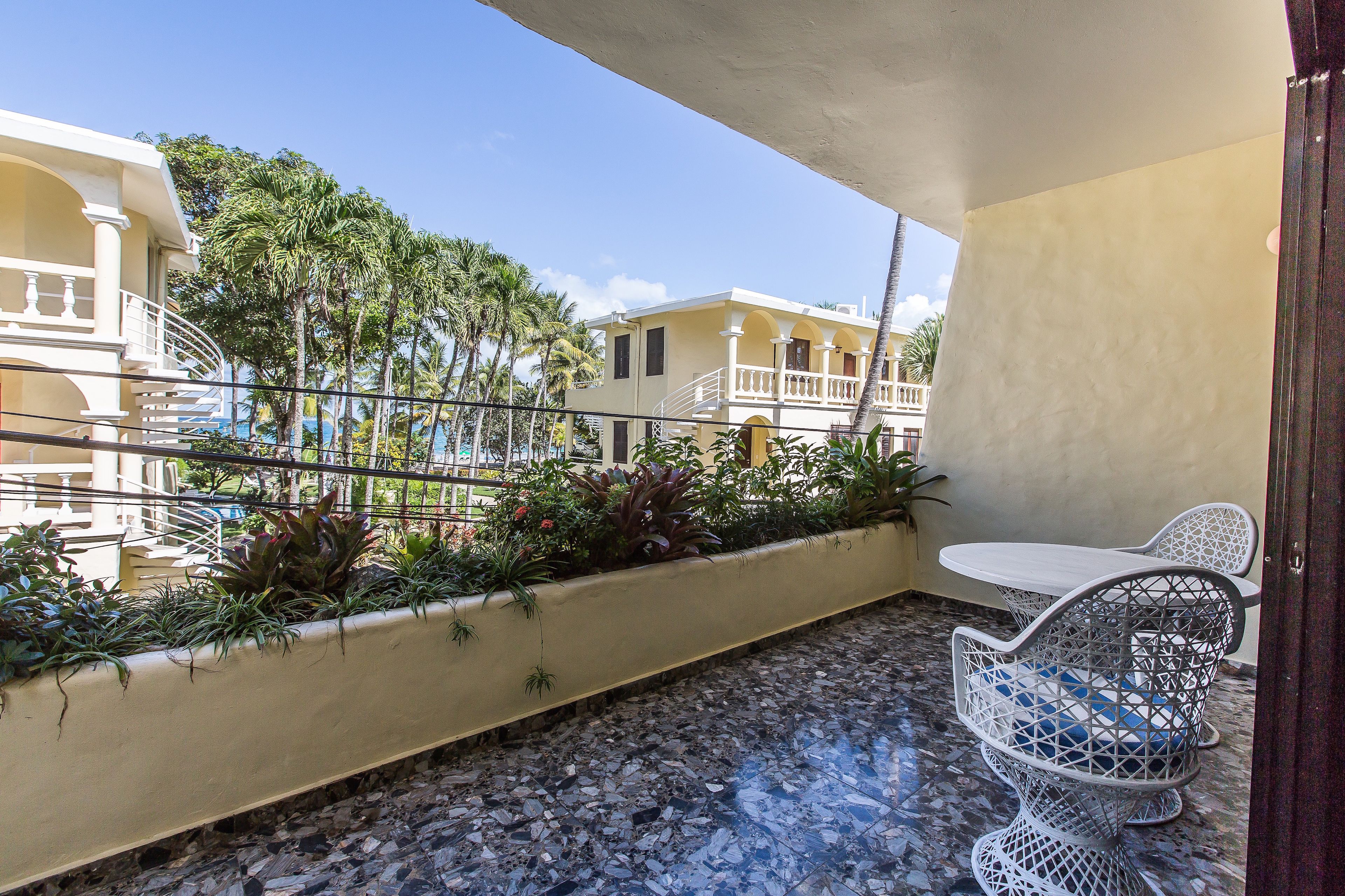 A balcony with a tiled floor, a white table, and a white chair. There are plants along the balcony edge, and buildings are visible in the background.